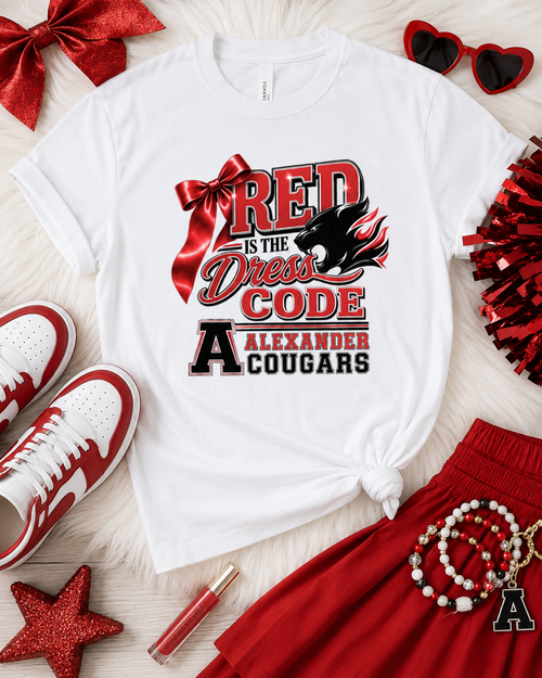 A white graphic T-shirt laid flat on a white faux-fur surface, styled with red and white sneakers, red pom-poms, red heart sunglasses, a red glitter star, a red bow, and red pleated skirt pieces. The shirt graphic reads “RED is the Dress Code Alexander Cougars” with a large red ribbon and black cougar graphic.