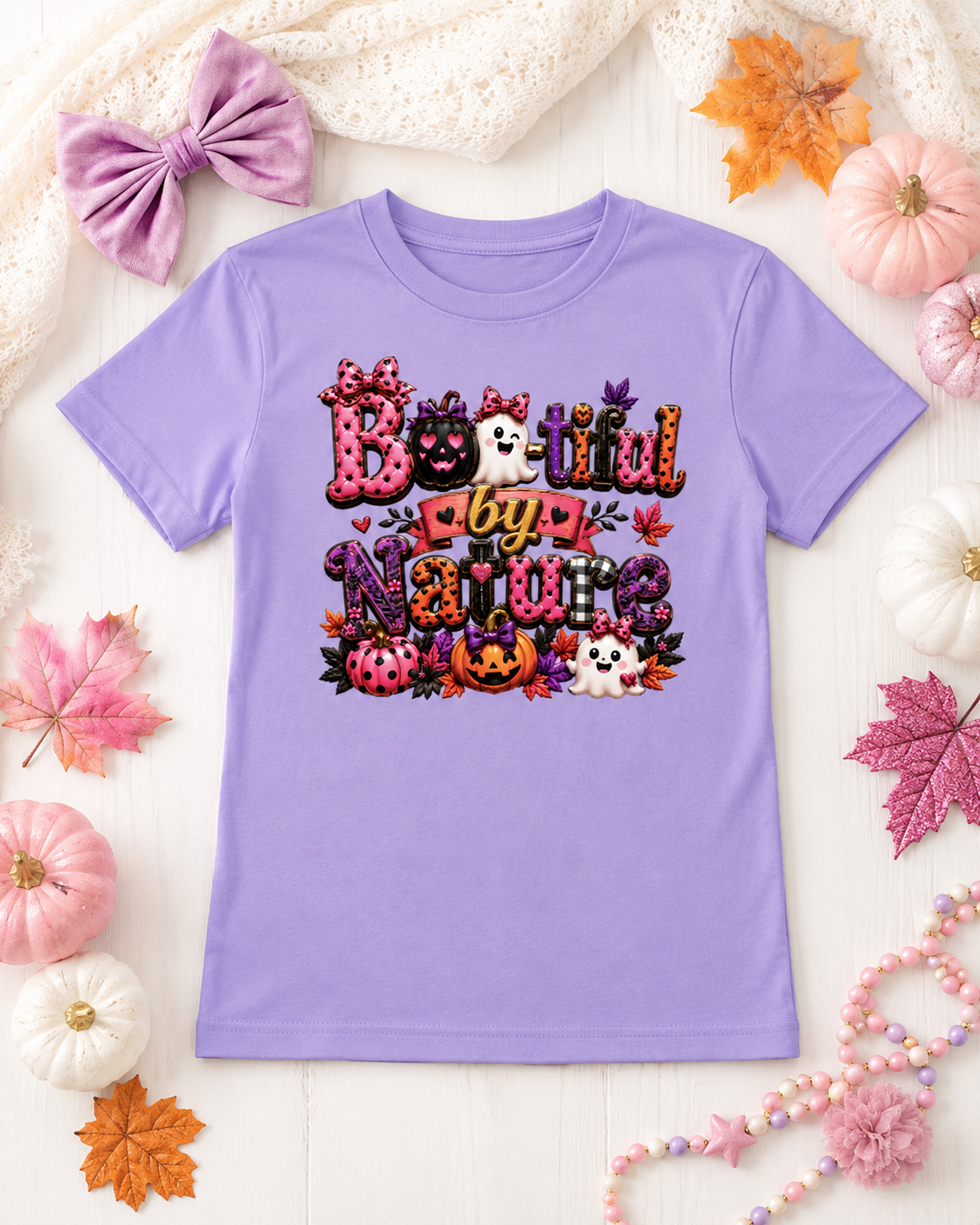 A lavender purple short-sleeve t-shirt with a colorful 'Booiful by Nature' Halloween graphic, styled in a flat lay with autumn leaves, pumpkins, and a lace bow on a white wood background.